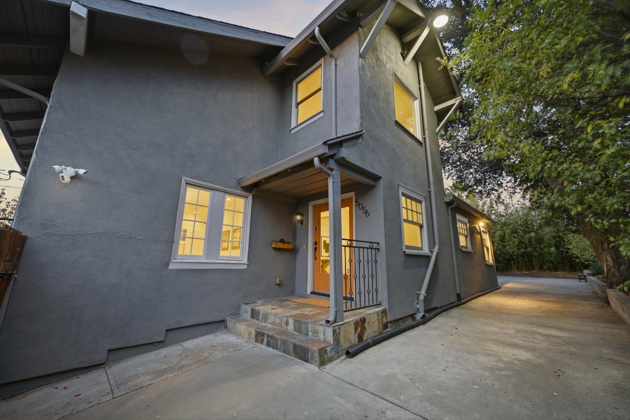 Two-story gray stucco house with warmly lit windows, a covered entrance, and a concrete driveway at dusk.
