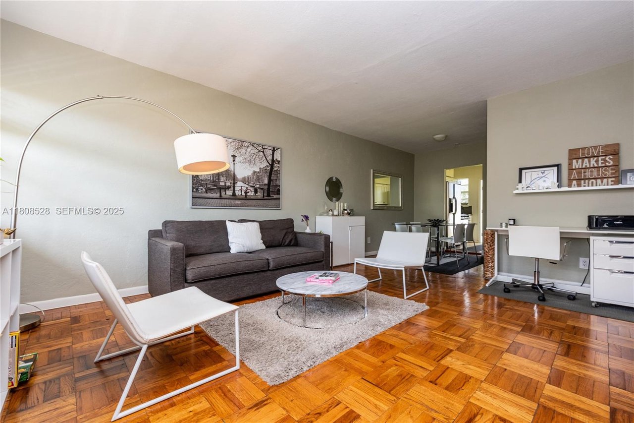 Furnished living room with parquet floors, gray sofa, white chairs, and an open dining area.