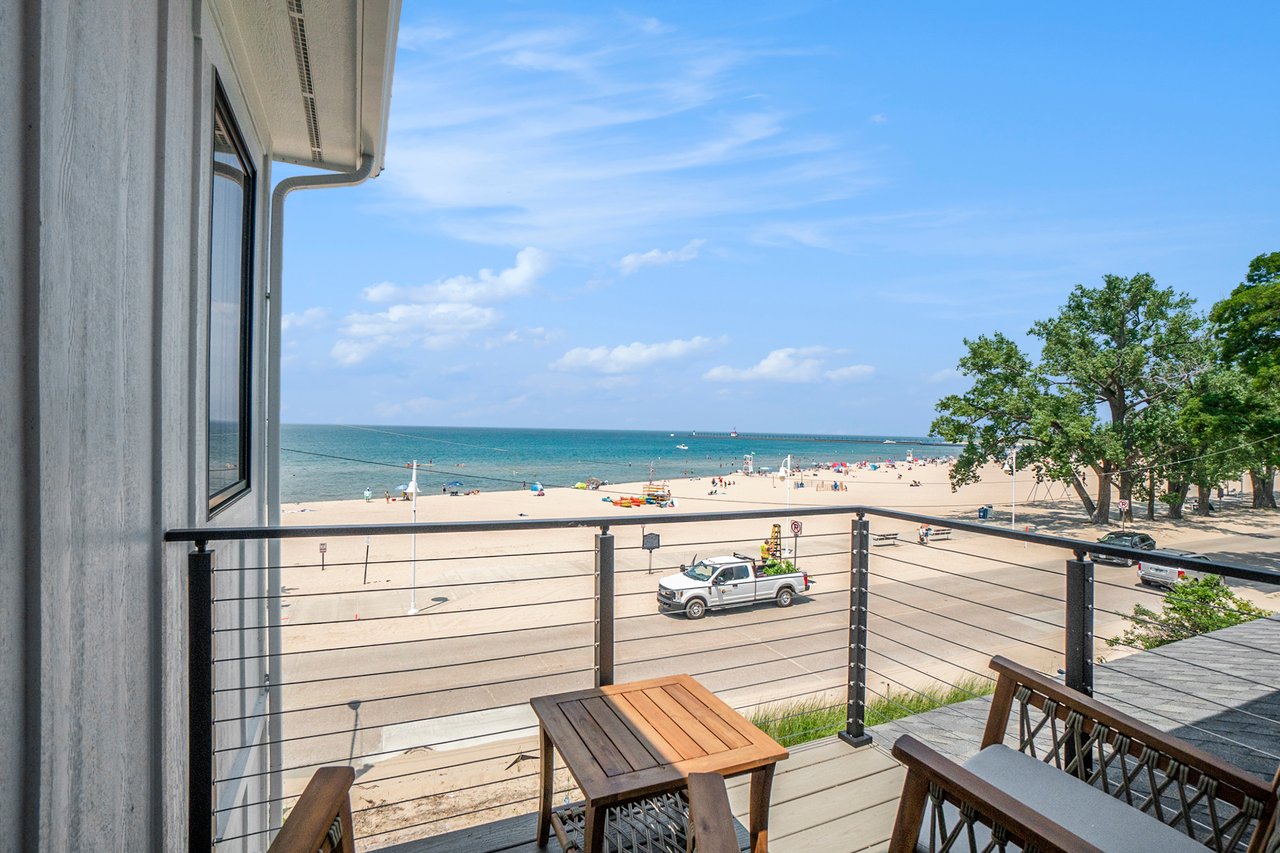 Balcony overlooking a sandy beach and blue water, with outdoor seating and a cable railing.