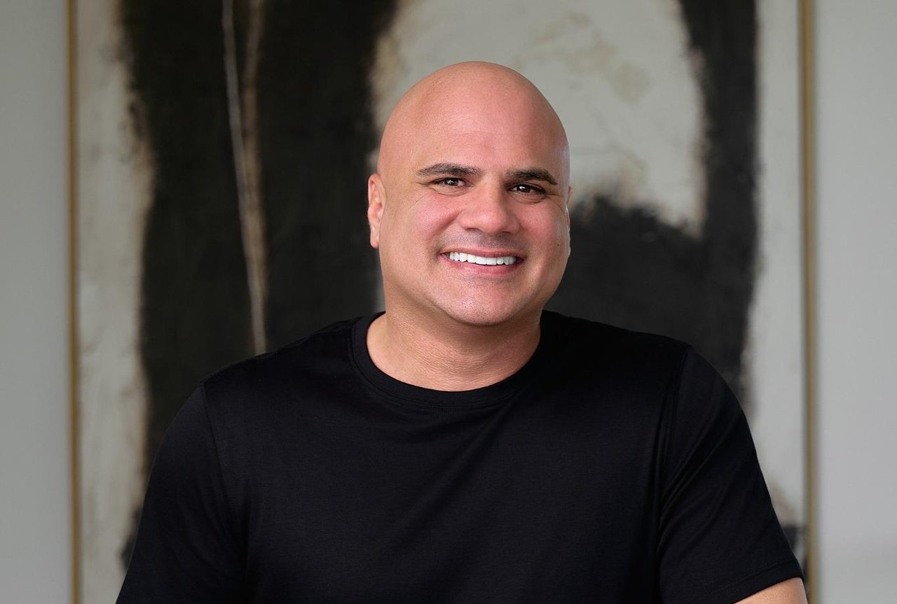Marc Coriaty smiling in a black shirt in front of abstract artwork.