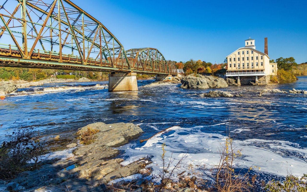 Steel truss bridge over the Androscoggin River rapids beside a historic mill in Topsham.