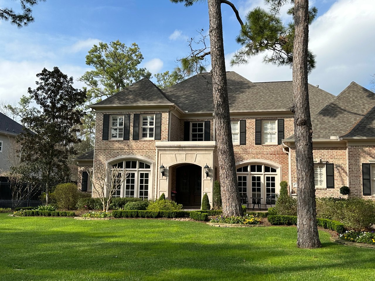 A Traditional two story luxury estate brick home with limestone entrance. It is landscaped with boxwood shrubs, green lawn, magnolia and tall pine trees. 