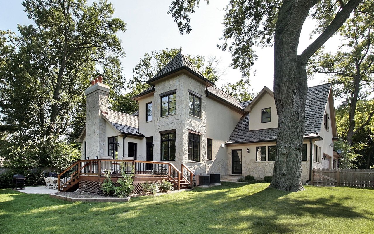 Two-story stone-and-stucco home with a raised deck, mature trees, and a broad lawn.