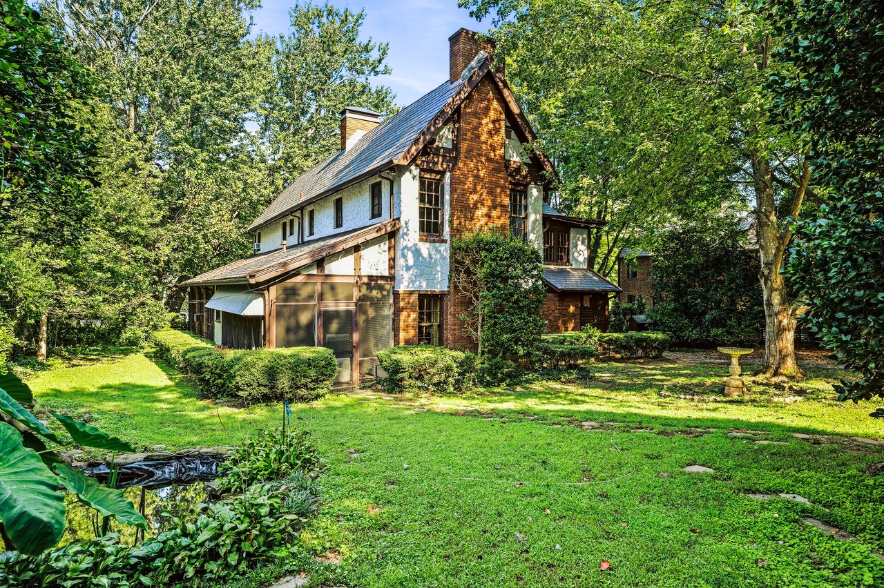 Two-story brick and white-sided house with steep gable, surrounded by mature trees and a landscaped lawn.
