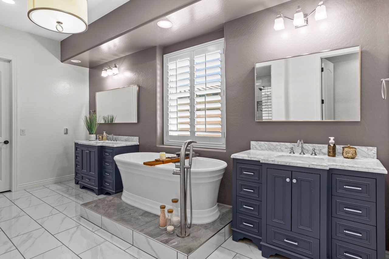 A spacious bathroom with a freestanding tub centered between two navy double-sink vanities, large mirrors, window shutters, and marble-look tile floors.