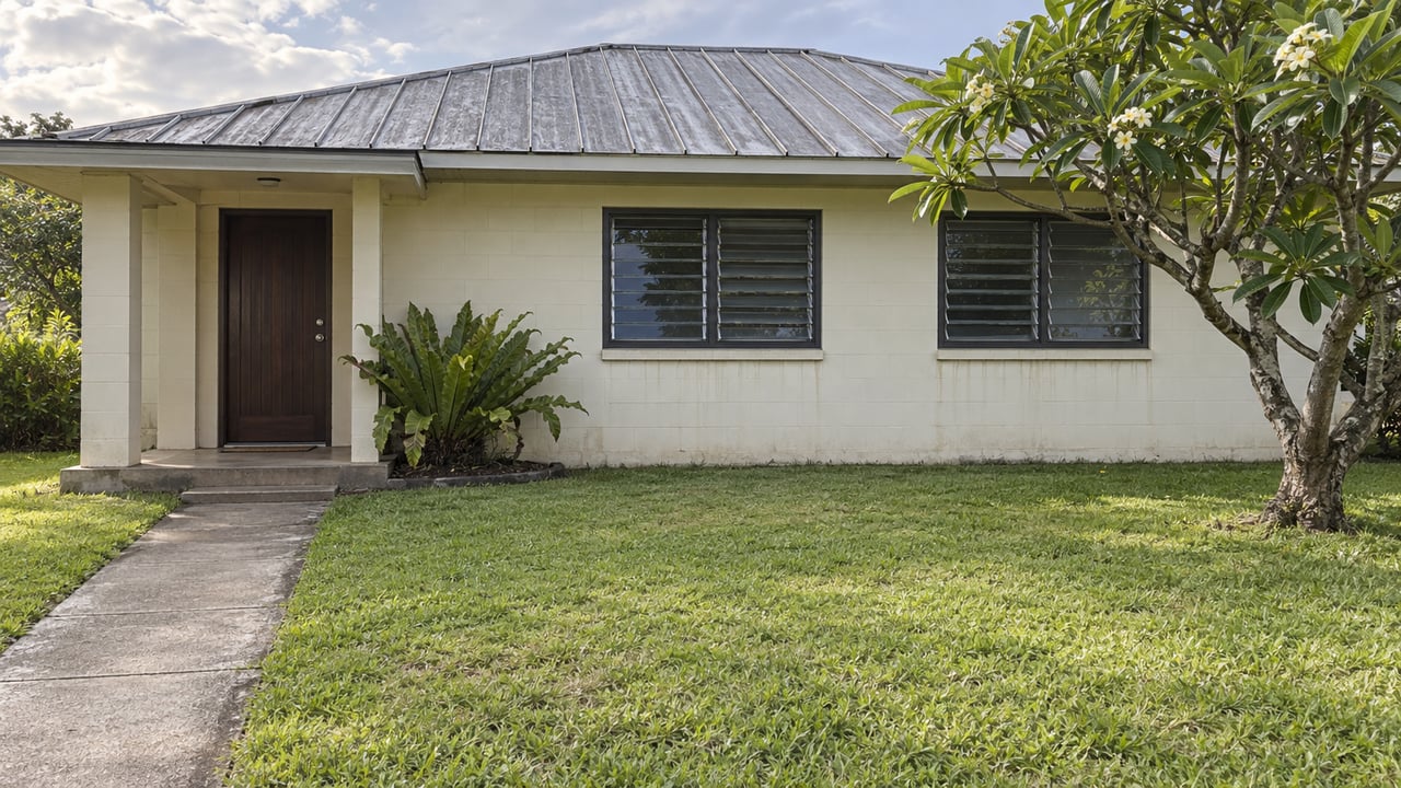 Older one-story house with a weathered metal roof, concrete approach, and plumeria tree in the front yard.