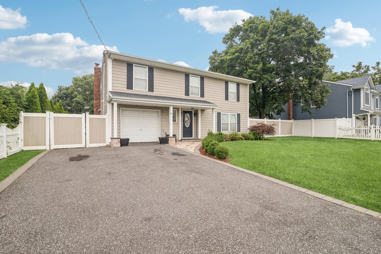 Two-story beige house with dark shutters, a wide driveway, and a fenced green yard.