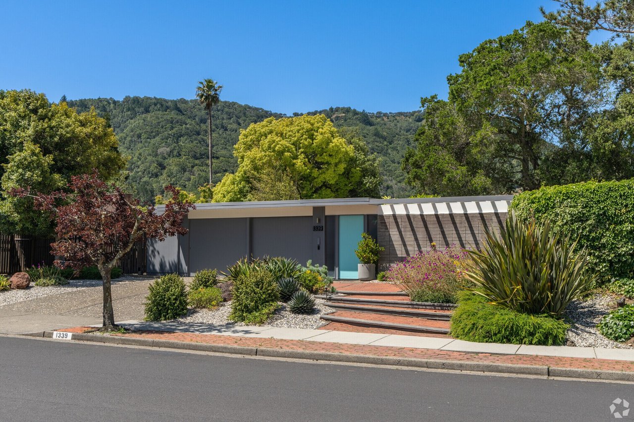 Modern single-story home with a turquoise front door, landscaped yard, and wooded hills behind it.