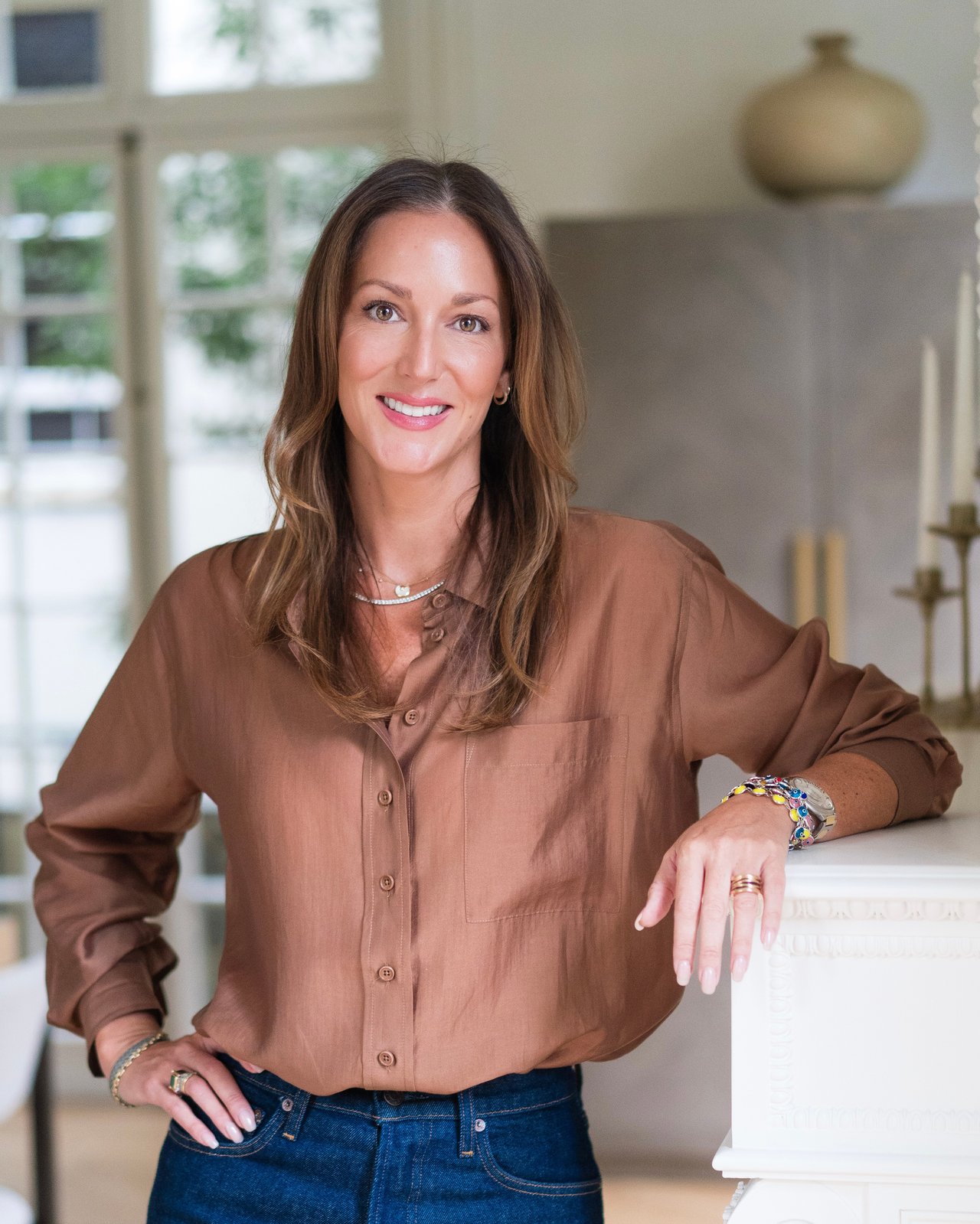 Samantha Miller poses beside a white mantel, wearing a brown blouse and colorful bracelets.