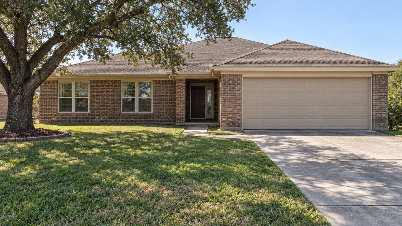 A single-story red-brown brick home with a broad front lawn, mature live oak, concrete driveway and tan two-car garage in clear daylight.