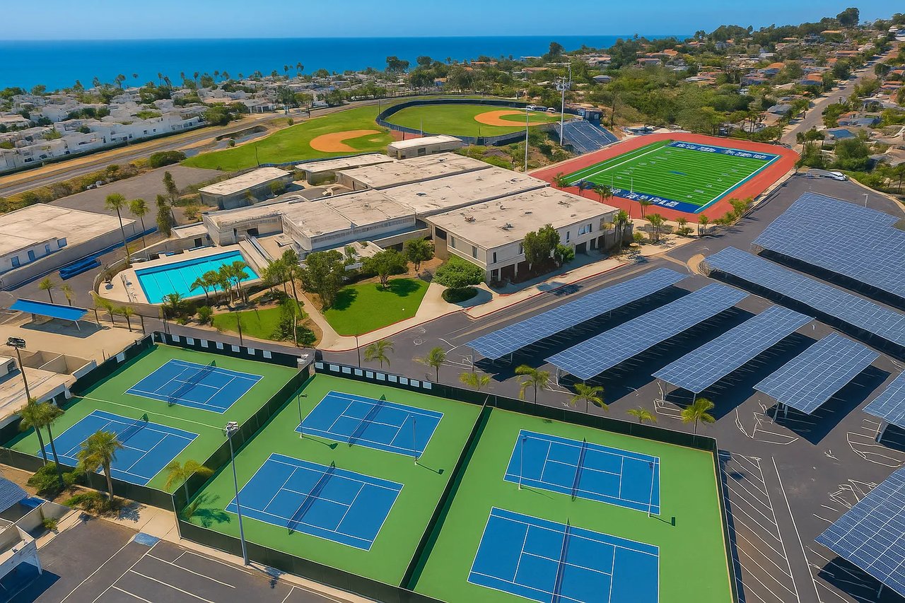 A bright aerial view of Dana Hills High School in Dana Point, showing tennis courts, swimming pool, solar-covered parking, football stadium, and baseball fields with the Pacific Ocean in the background.