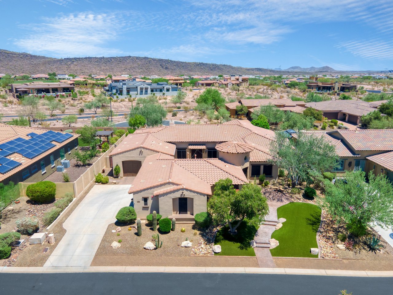 Aerial view of a suburban home with a tile roof, driveway, desert landscaping, and neighboring houses in a dry hillside community.