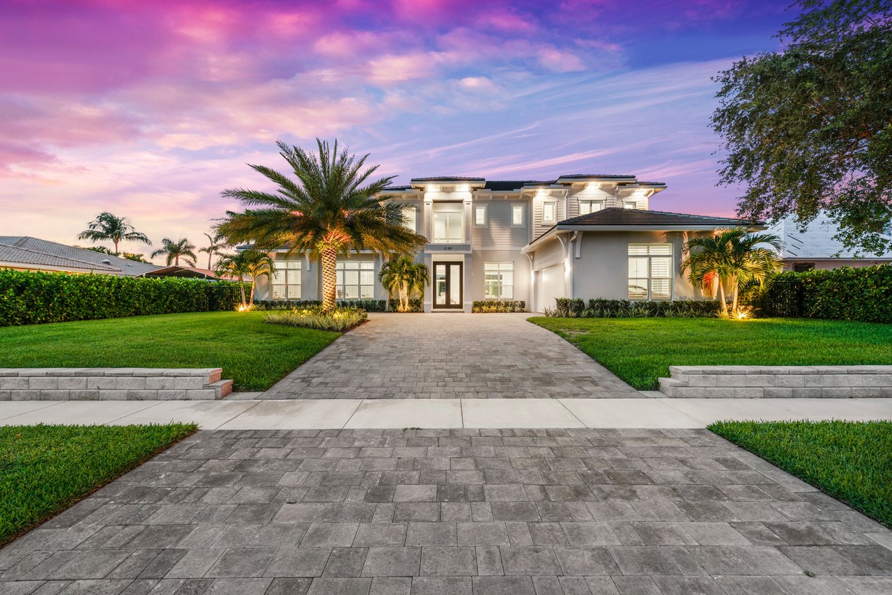Modern two-story residence with a wide paved driveway and palm trees at dusk.