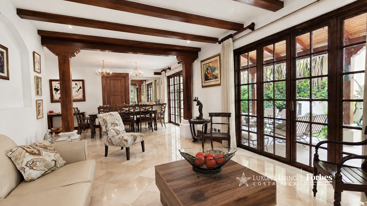 Living and dining room with dark wood ceiling beams and French doors opening to a leafy patio.