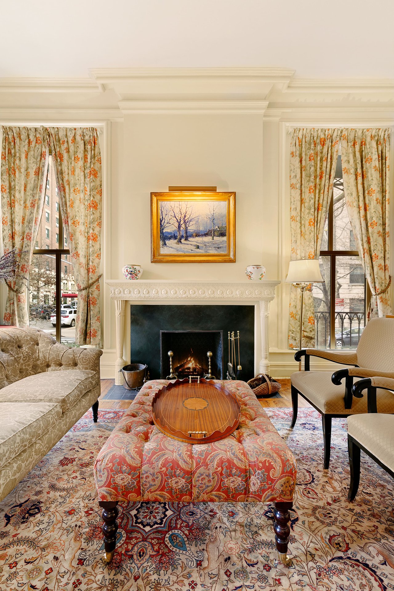 Elegant living room centered around a patterned red ottoman with a wooden tray, facing a classic fireplace and tall windows with floral curtains.