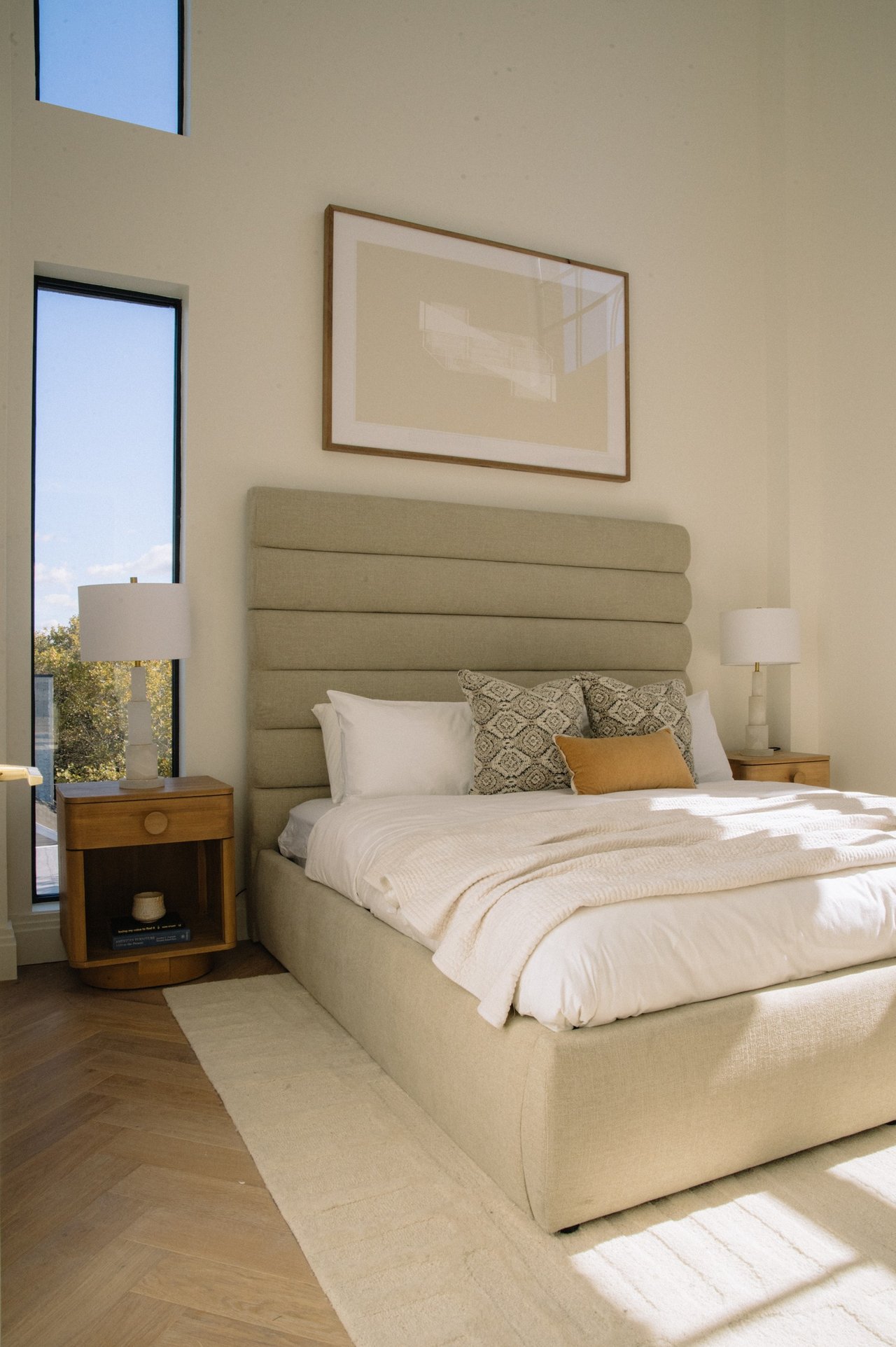 Furnished bedroom with a tall beige headboard, white bedding, patterned pillows, and sunlight through tall windows.