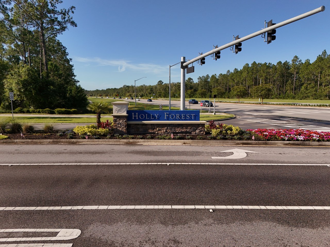 Holly Forest entrance sign beside a landscaped intersection and pine trees.