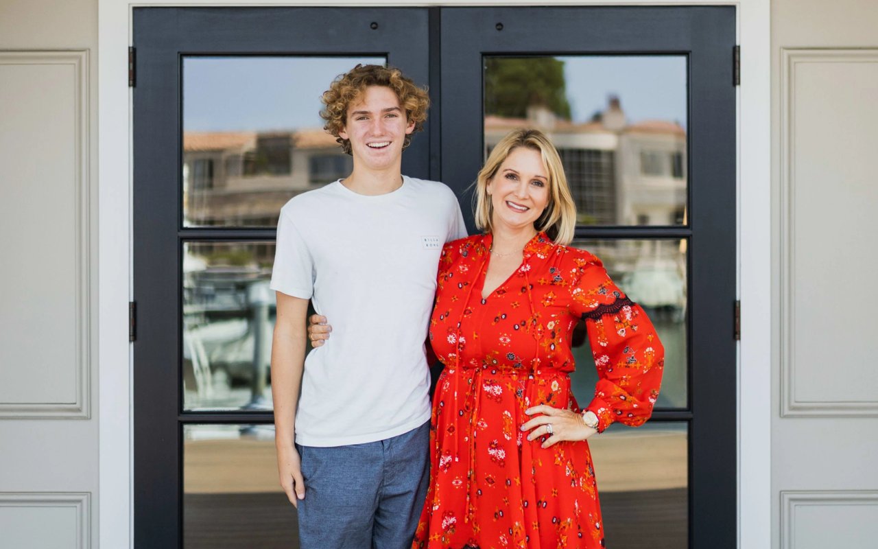 A mother in a red floral dress and her teenage son in a white t-shirt stand together, smiling in front of glass doors.