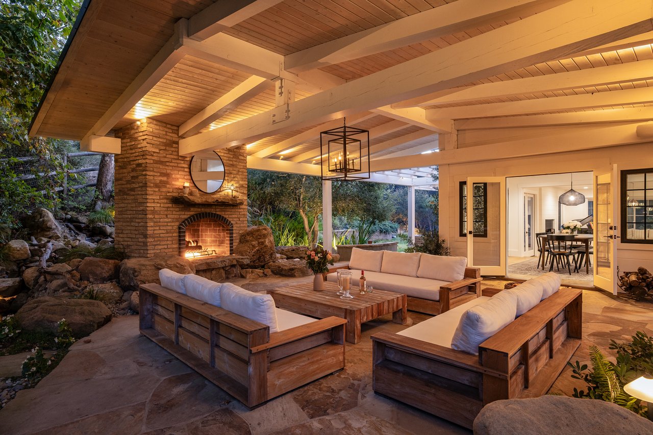 Covered patio with a stone fireplace, wood lounge seating, and a rocky wooded garden.