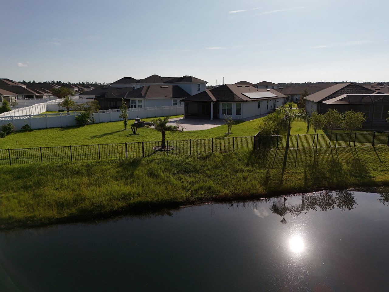 Aerial view of neighborhood homes beside a pond, with fenced grassy backyards.