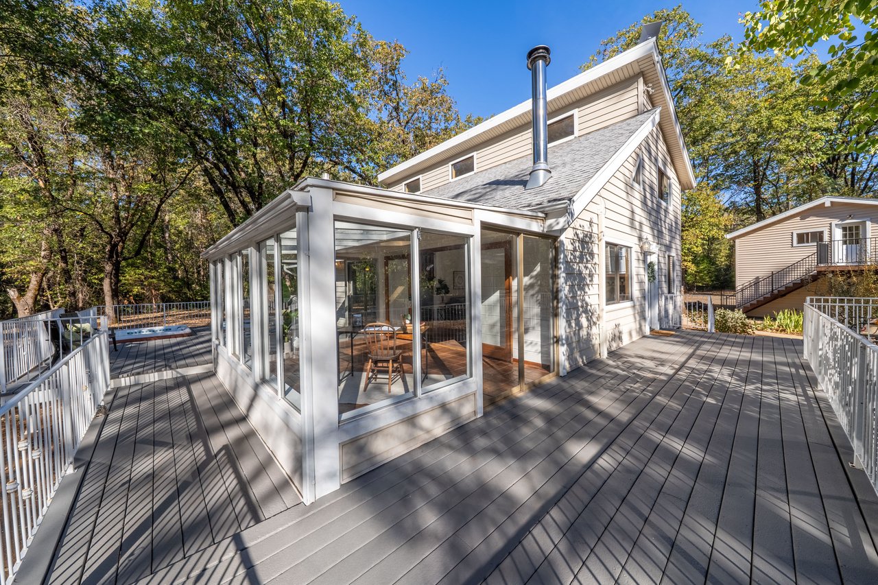 Two-story home with a glass-enclosed sunroom, expansive deck, and hot tub among autumn trees.