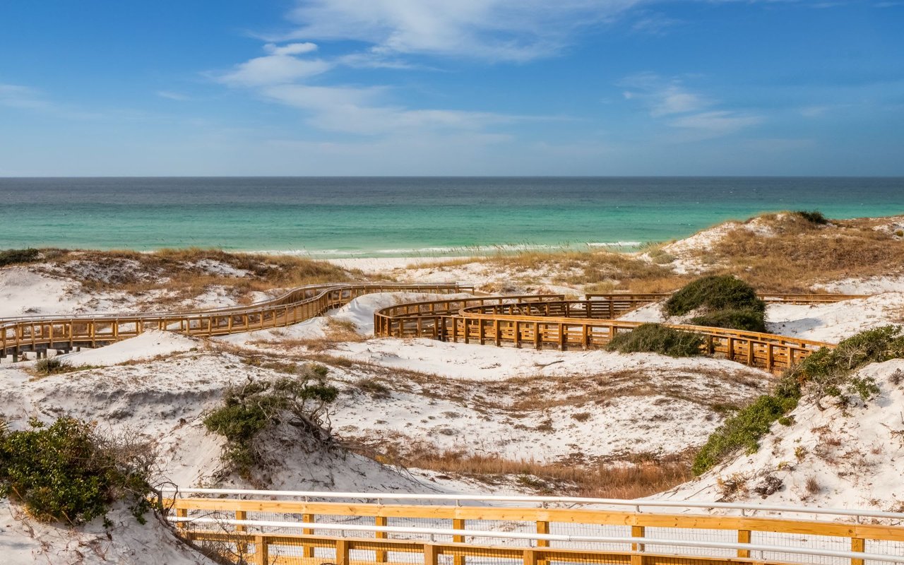 A curved wooden boardwalk winds through sandy dunes and sea oats, leading to a white-sand beach.