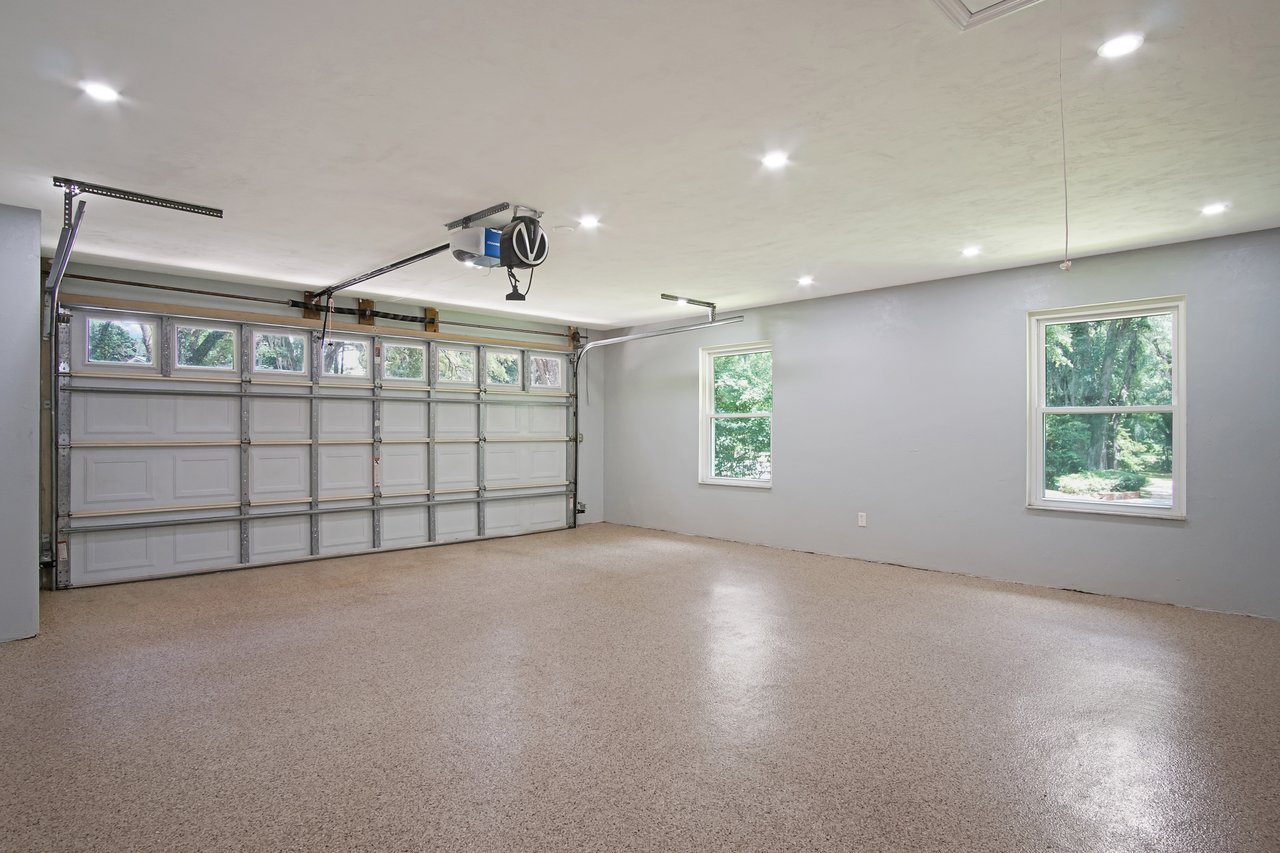 Spacious, well-lit garage interior with beige epoxy flooring, white walls, large closed door, and two windows showcasing greenery outside. Minimalist and clean.