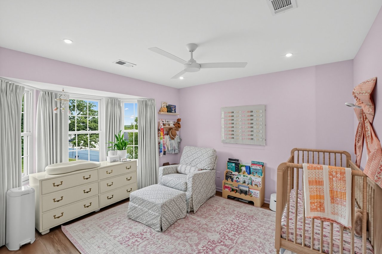 Nursery with a wooden crib, lavender walls, and a patterned armchair beside tall windows.