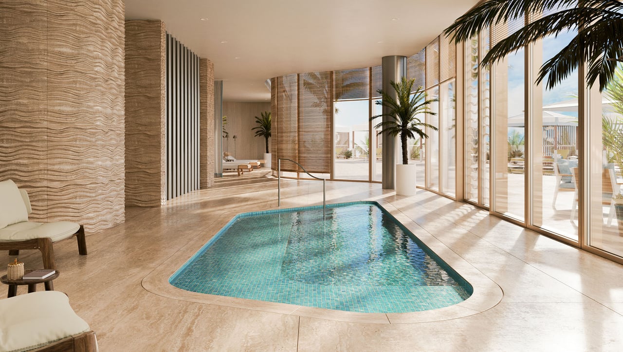 Indoor pool with turquoise tile, floor-to-ceiling glass doors, palm trees, and lounge seating.