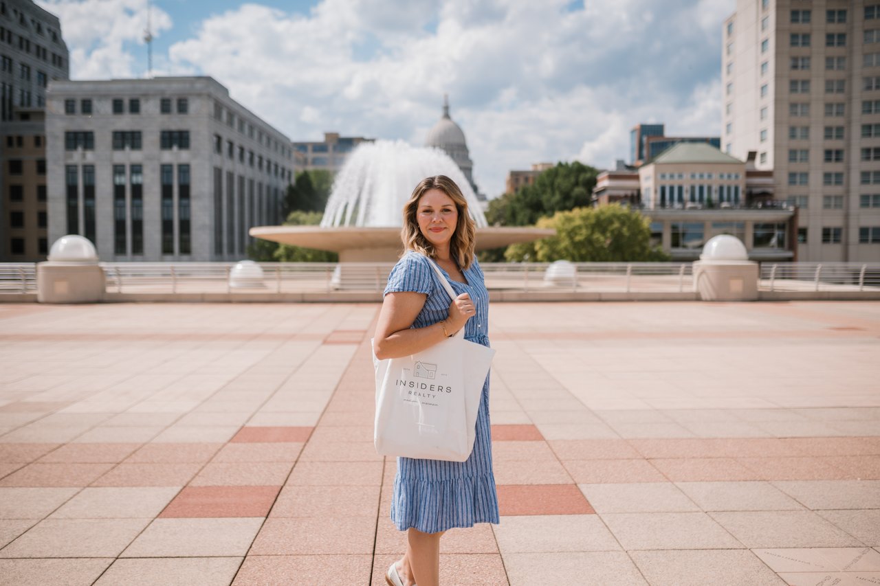 Cait Berry of Insiders Realty standing on the Capitol Square in downtown Madison on a bright summer day, with the fountain and State Capitol dome behind her, carrying an Insiders Realty tote.