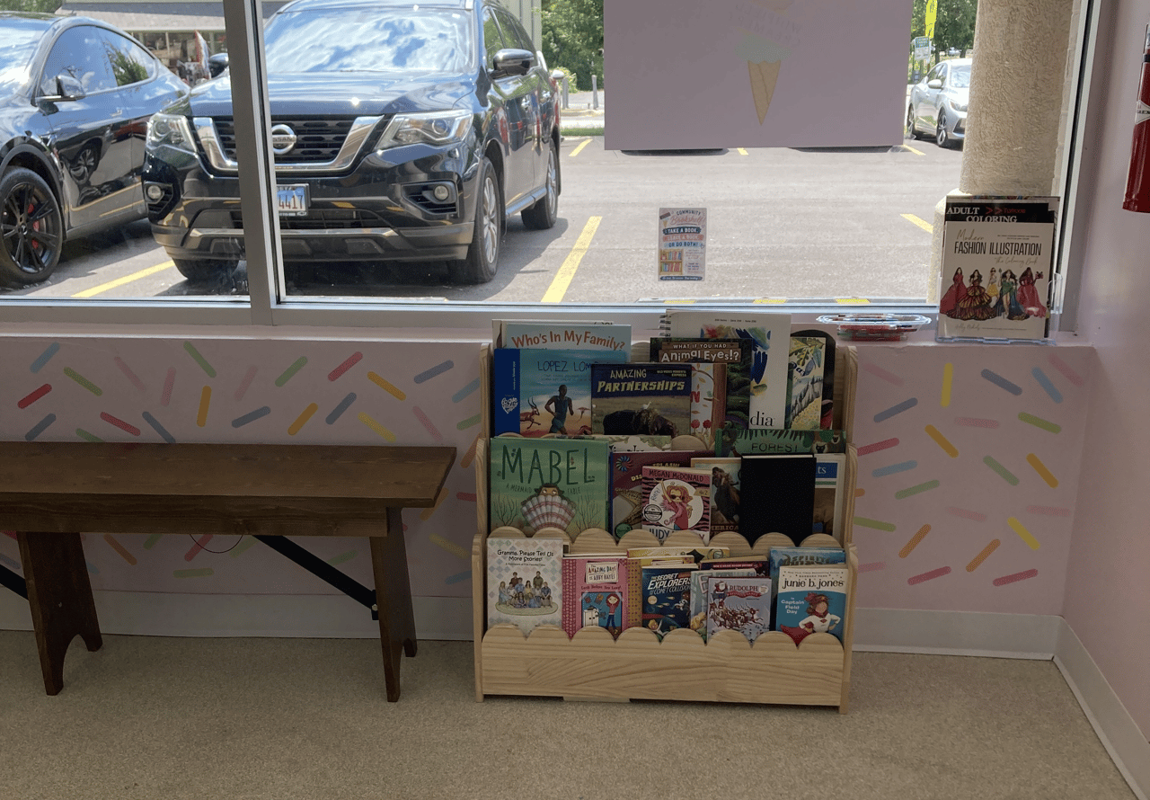 A light wooden children's bookshelf filled with books sits beneath a window with sprinkled wallpaper in the Winfield Creamery ice cream shop