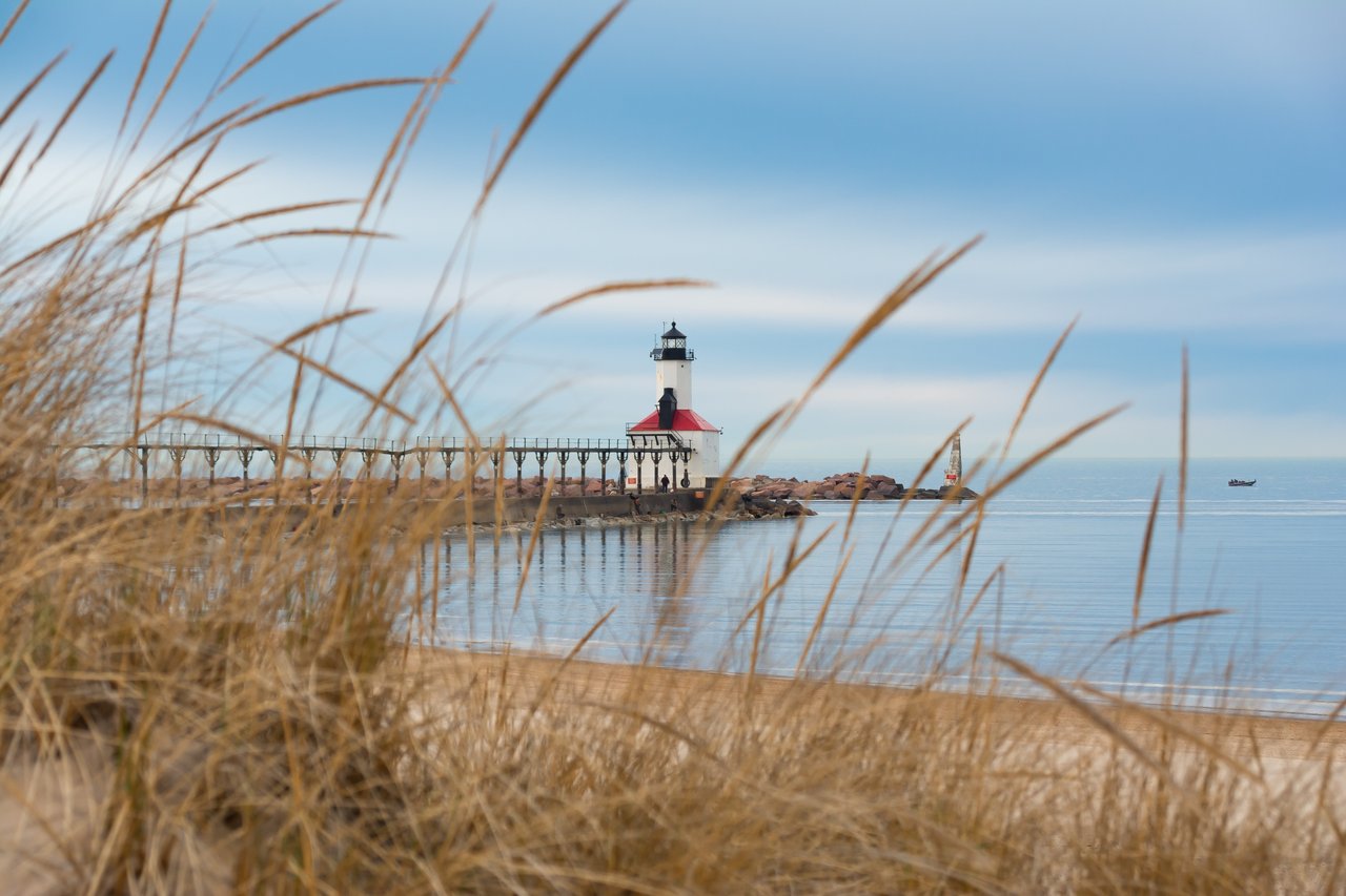 White lighthouse with a red roof on a rocky pier, framed by beach grasses beside calm water.