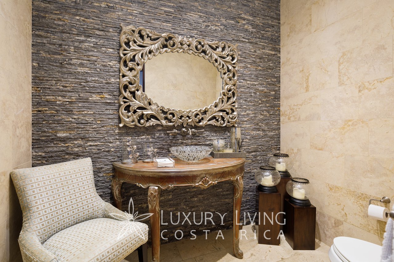 Bathroom with an ornate mirror, carved console table, patterned chair, and dark stacked-stone accent wall.