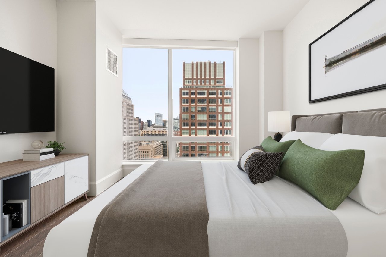 Furnished bedroom with green pillows and large windows overlooking downtown buildings.
