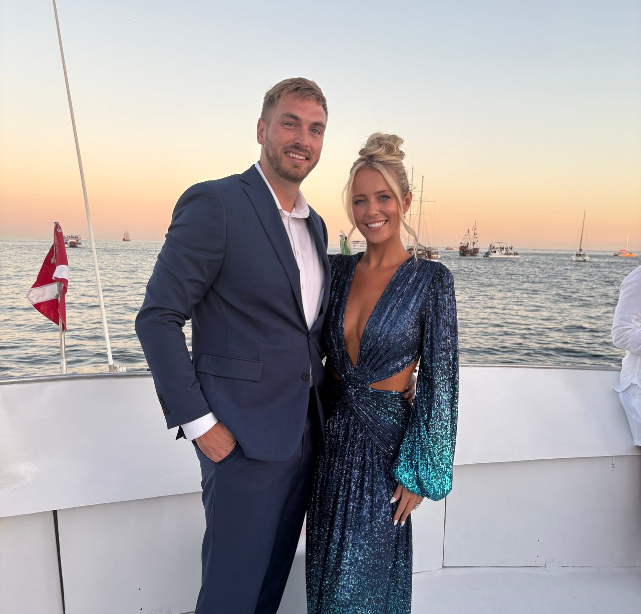 A man in a navy suit and a woman in a sparkling blue dress pose on a boat at sunset.