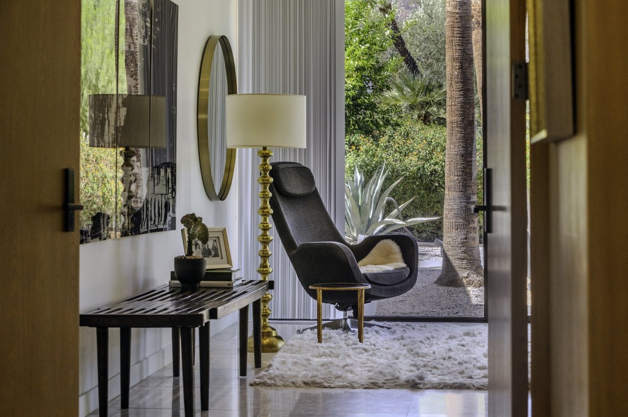 Modern sitting area with a charcoal lounge chair facing a glass door to a palm-filled garden.