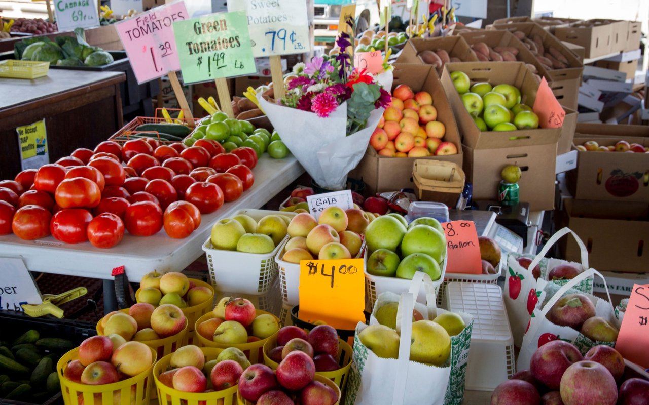 Farmers market produce stand with red tomatoes, apples, green tomatoes, and handwritten price signs.