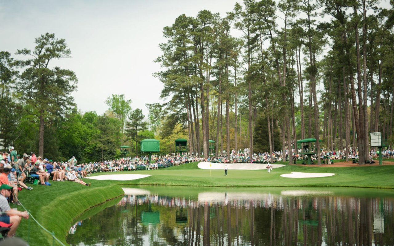 Golfers on a tree-lined course as spectators watch from beside a pond.