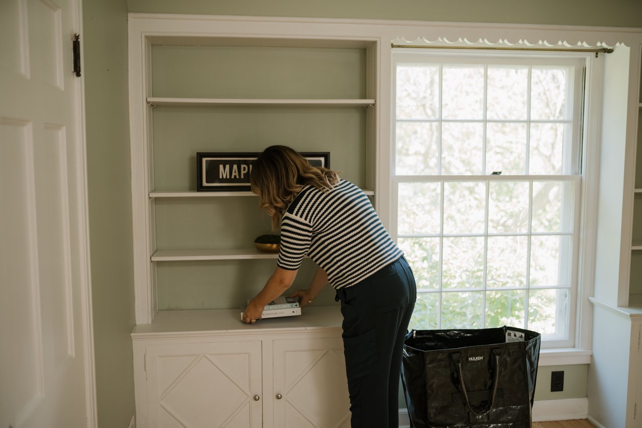 A person clearing books and keepsakes from a built-in shelf while downsizing an older home's belongings.