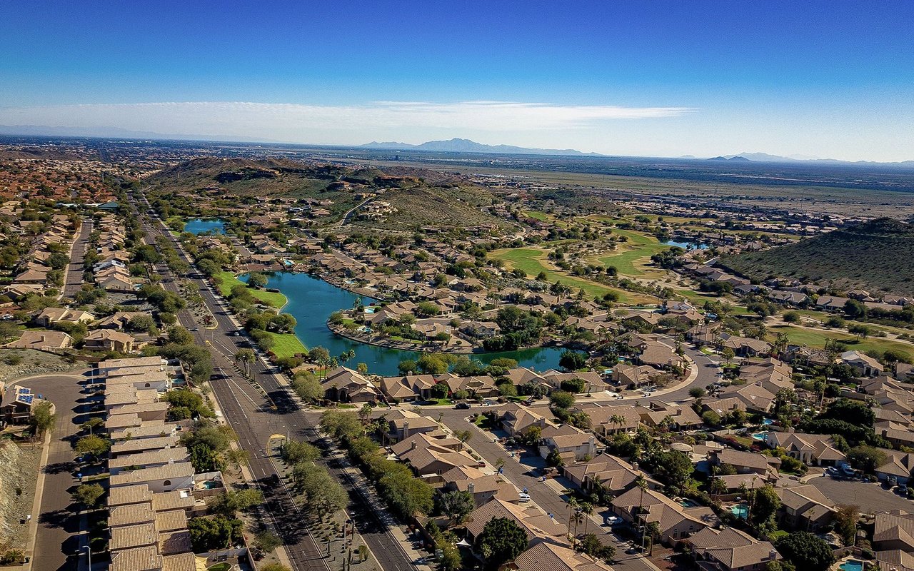 Aerial view of a desert-suburban neighborhood with lakes, a golf course, and distant mountains.
