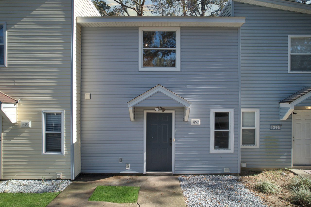 Exterior of a modern townhome with blue-gray siding, a central dark door, and two windows. A small green mat and gravel landscaping enhance its neat look.