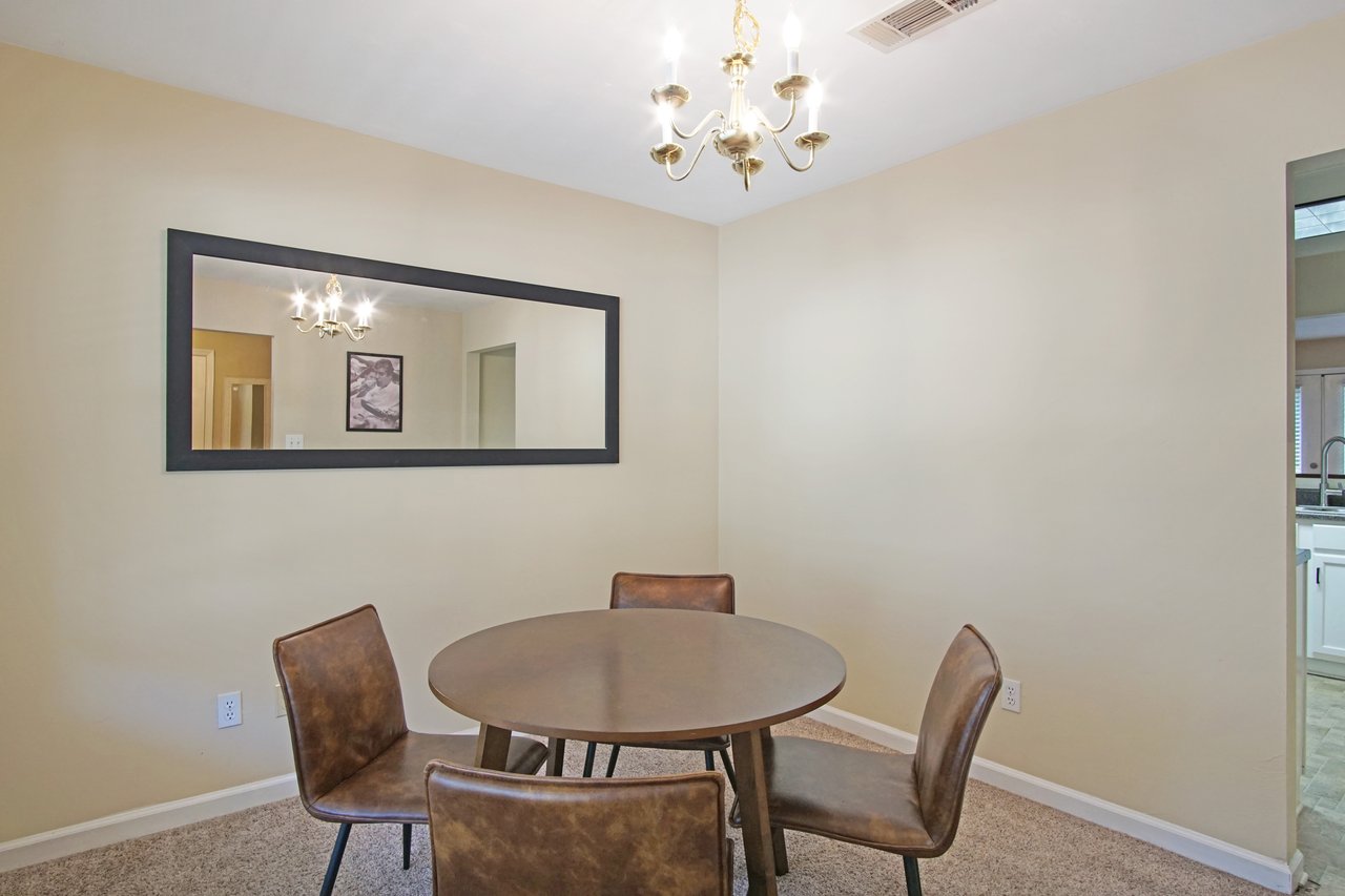 Dining area at 529 Collinsford Road, Tallahassee, Florida, featuring a round wooden table with four brown leather chairs, a large wall mirror, and a gold chandelier. The space connects to the kitchen through an open doorway and maintains a neutral color palette with beige walls and carpet flooring.