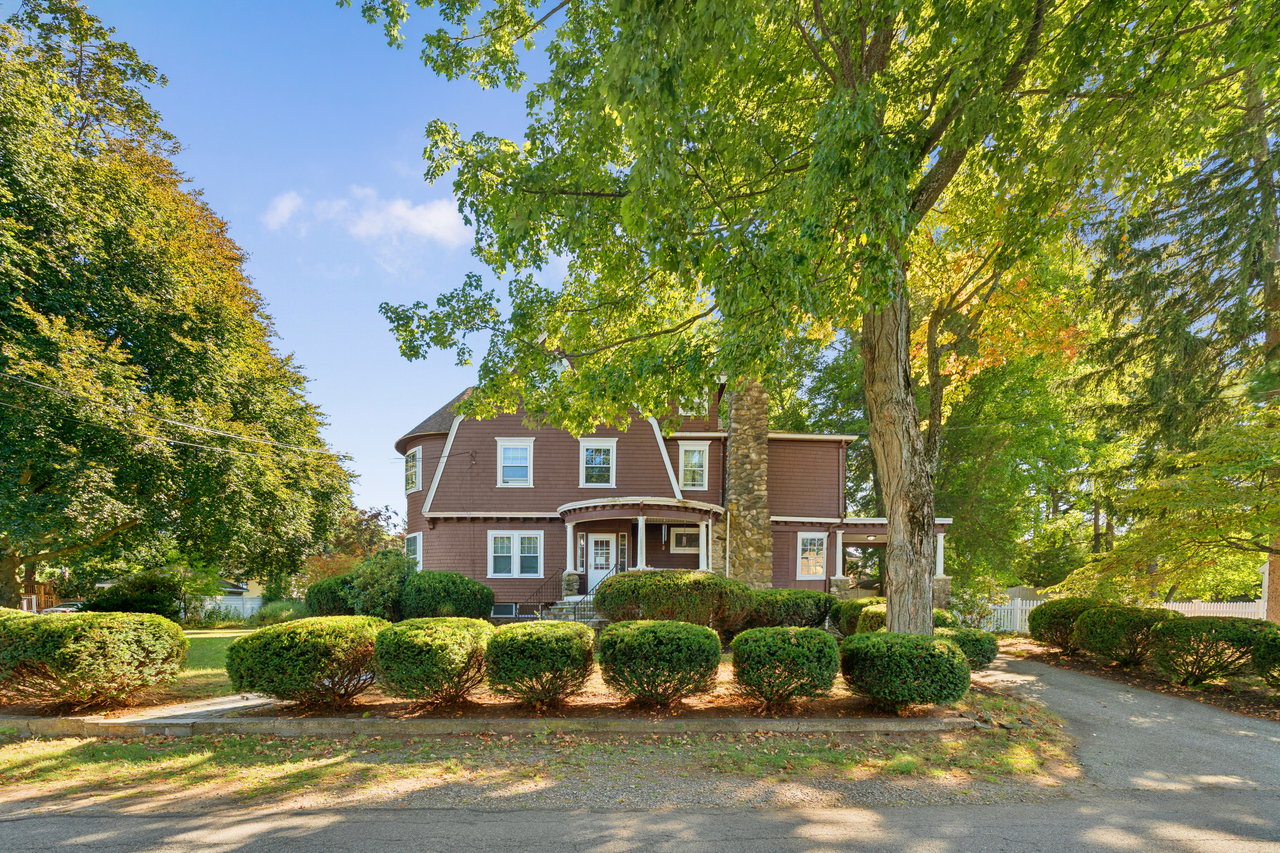 Brown-shingle house with a stone chimney, curved porch, and mature trees.