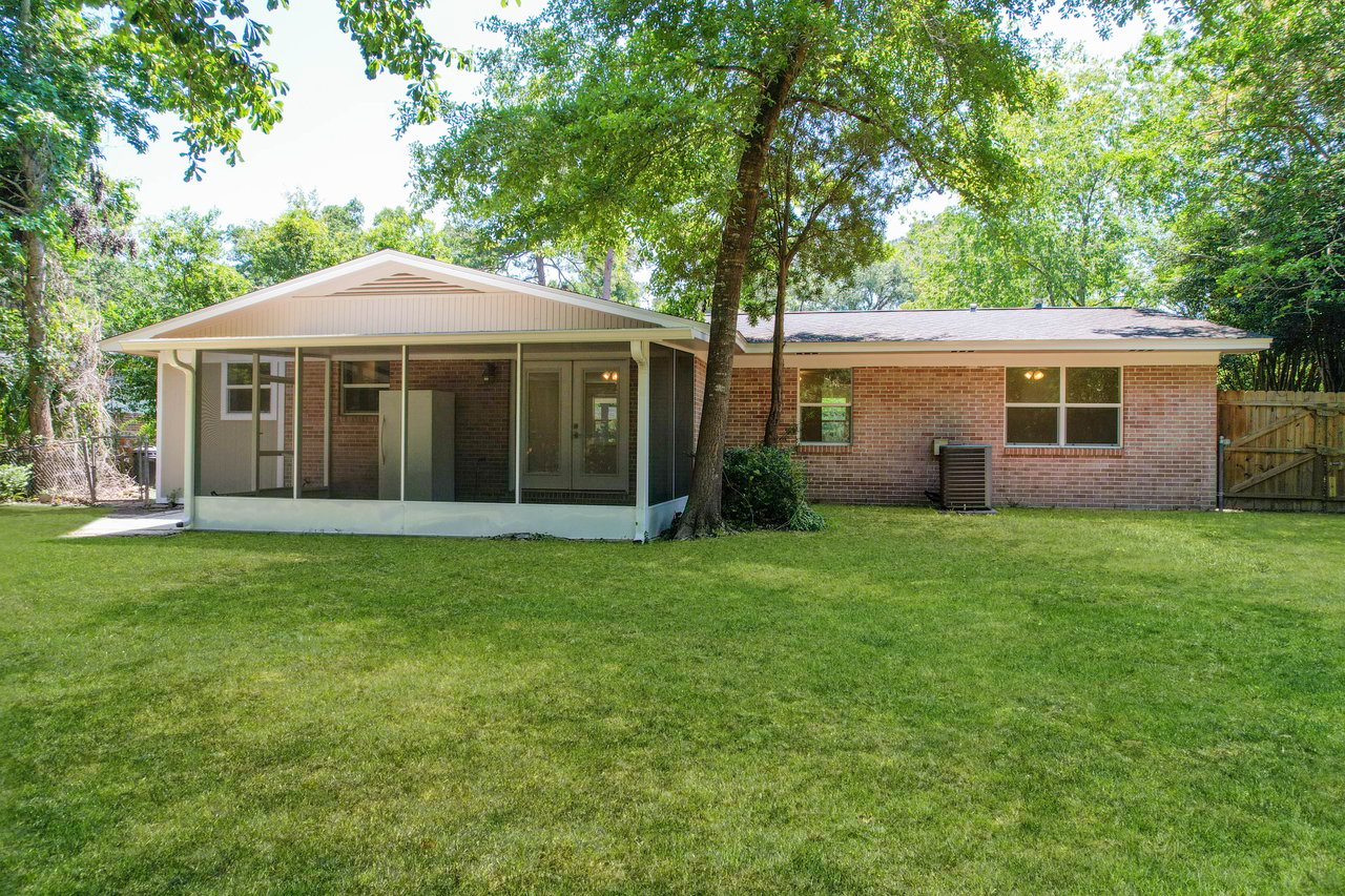 Brick house with a spacious backyard, shaded by trees. The patio has a screened porch and the setting is tranquil and lush with greenery.