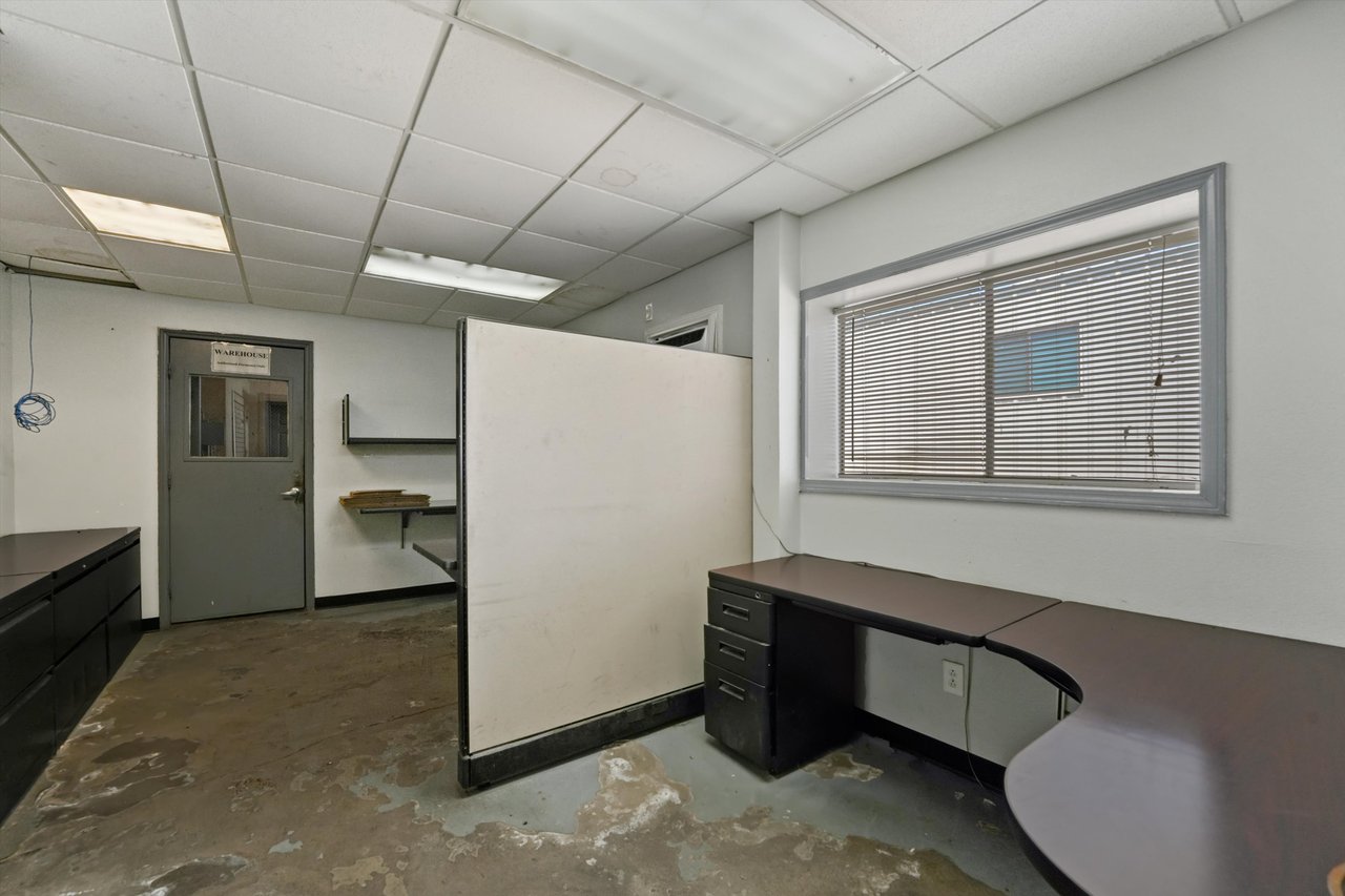 Warehouse office with dark desks, a central partition, and a door labeled “WAREHOUSE”.