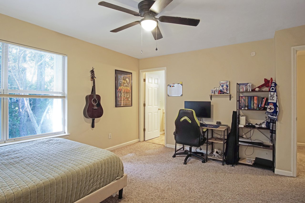 Bedroom at 529 Collinsford Road, Tallahassee, Florida, featuring beige walls, carpet flooring, and a ceiling fan with a light fixture. The room includes a bed, a desk with a computer setup, a black office chair, a wall-mounted guitar, and a bookshelf, creating a comfortable multipurpose space for rest and work.