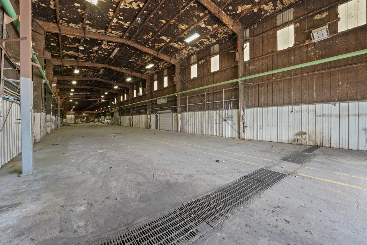 Vacant warehouse interior with high timber-truss ceilings, overhead piping, and floor drainage grates.