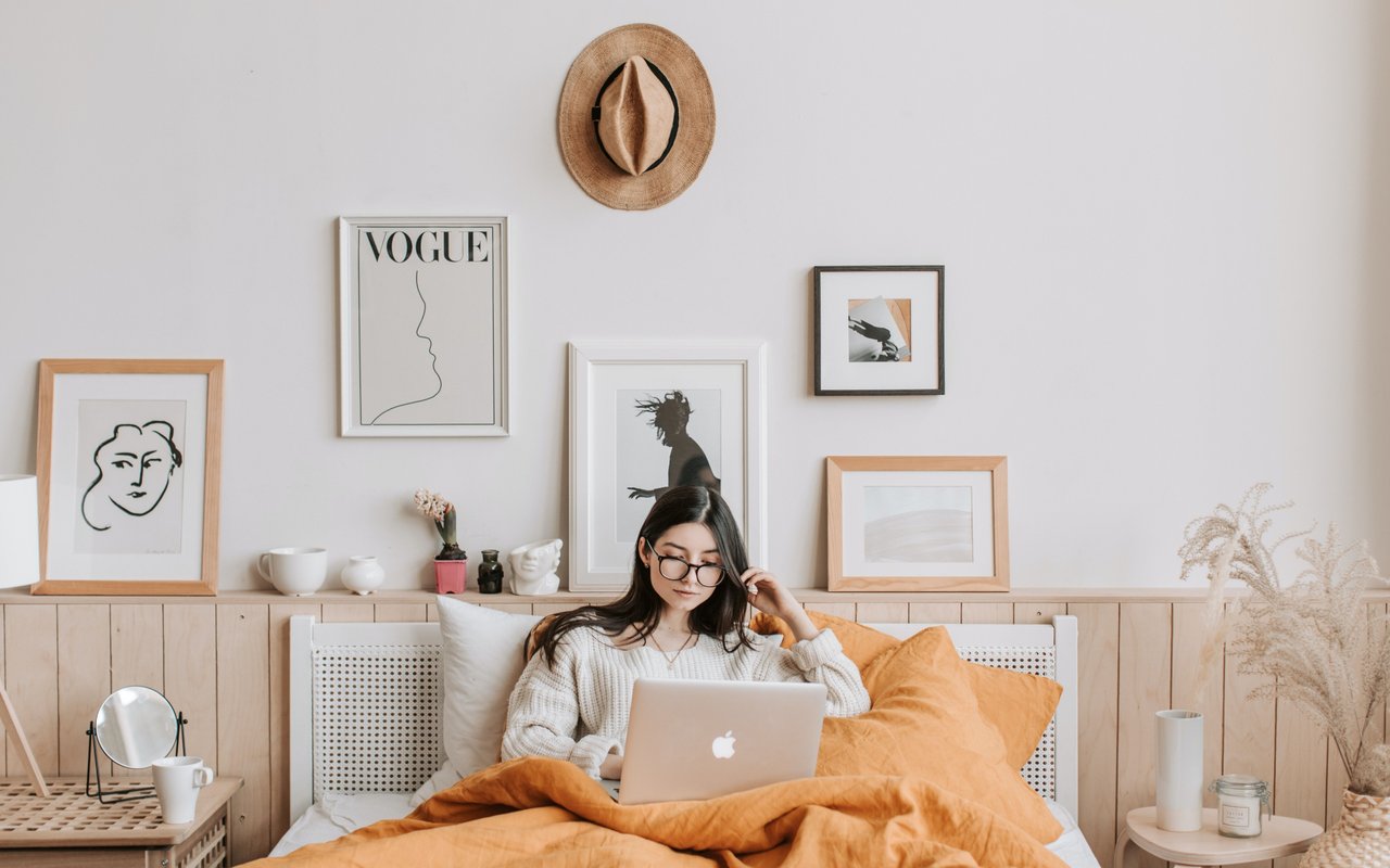 Woman sits in a cozy bed with orange bedding, working on a laptop, surrounded by framed art and minimal decor.