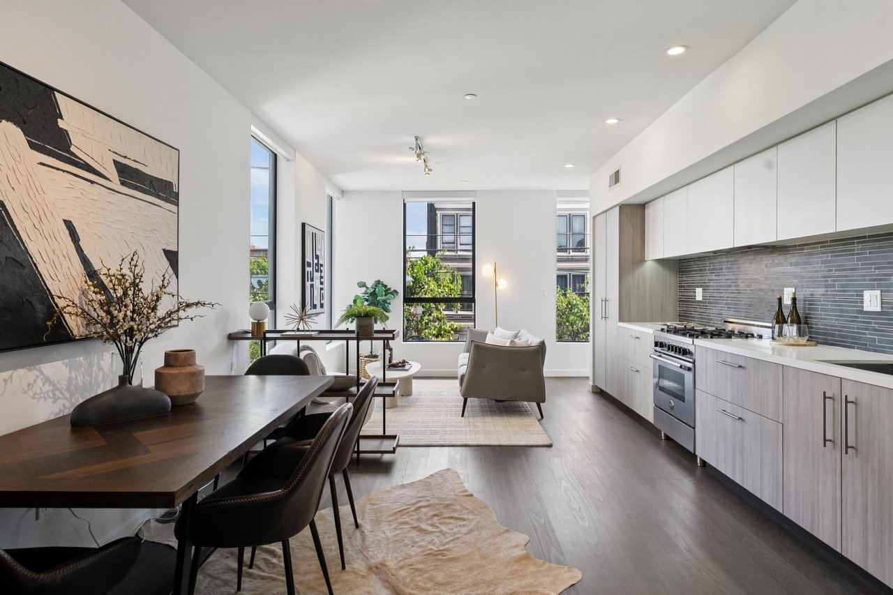 Open-plan apartment kitchen and living area with gray tile backsplash, wood dining table, and tall windows.