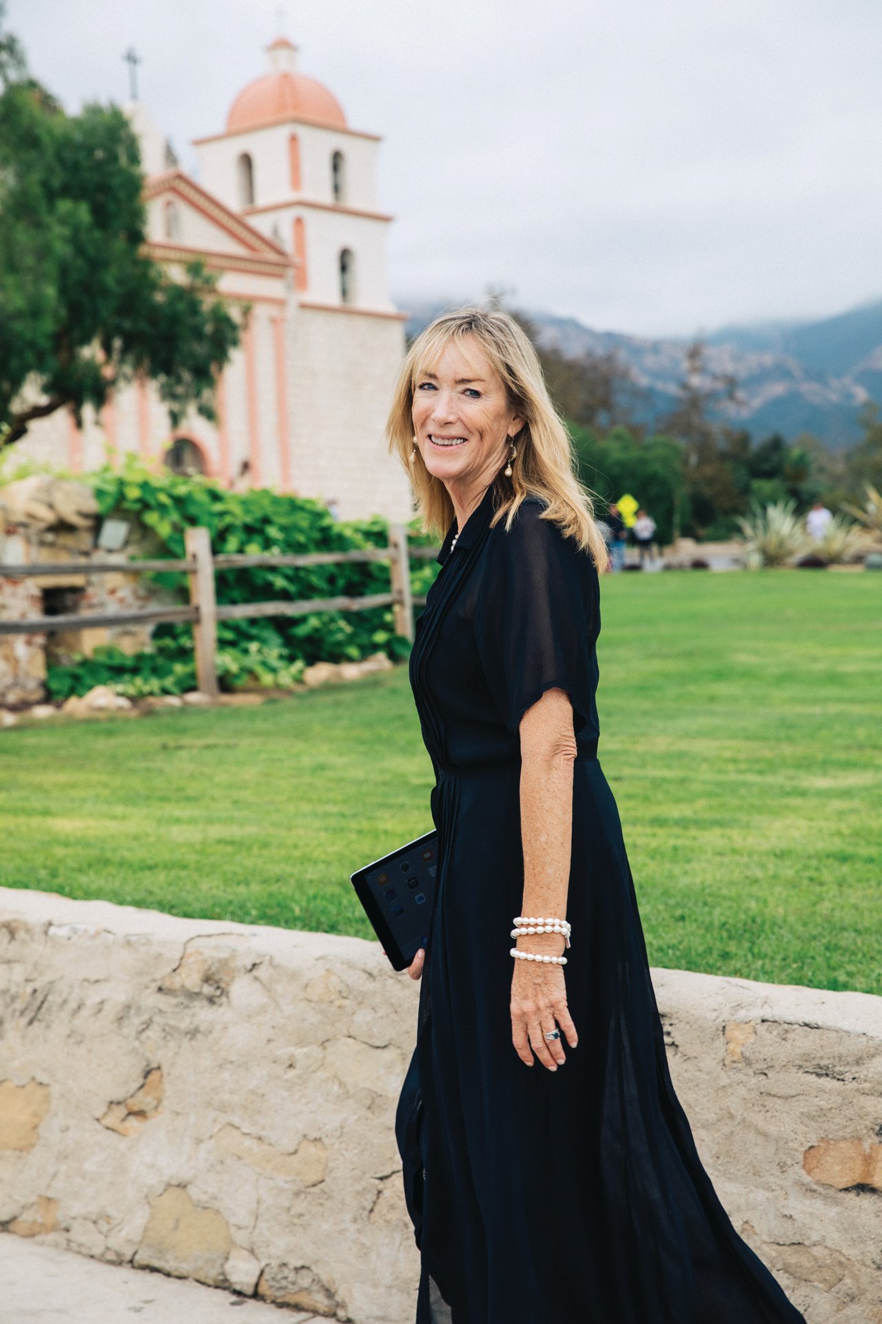Colleen Beall in a black dress holding a tablet on a lawn, with a church and mountains behind her.